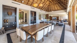 A spacious living area with a large wooden dining table, white chairs, a sofa, and a ceiling with a thatched roof design.
