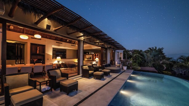 An infinity pool with a clear view of the sky, a covered outdoor seating area with lounge chairs, and a well-lit interior space with a bar and dining area.