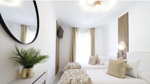 Habitación de hotel con una cama grande, una cama más pequeña, un espejo, un televisor, una planta y una lámpara decorativa.