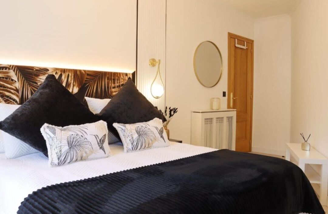Une chambre à coucher bien rangée, avec un lit, deux oreillers noirs, un oreiller blanc orné de motifs de feuilles, un plaid noir, une table de chevet avec une lampe, une petite table blanche avec un vase, et un miroir rond accroché au mur.