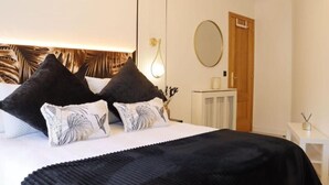 Un dormitorio ordenado con una cama, dos almohadas negras, una almohada blanca con diseño de hoja, un plaid negro, una mesita de noche con una lámpara, una pequeña mesa blanca con un florero y un espejo redondo en la pared.