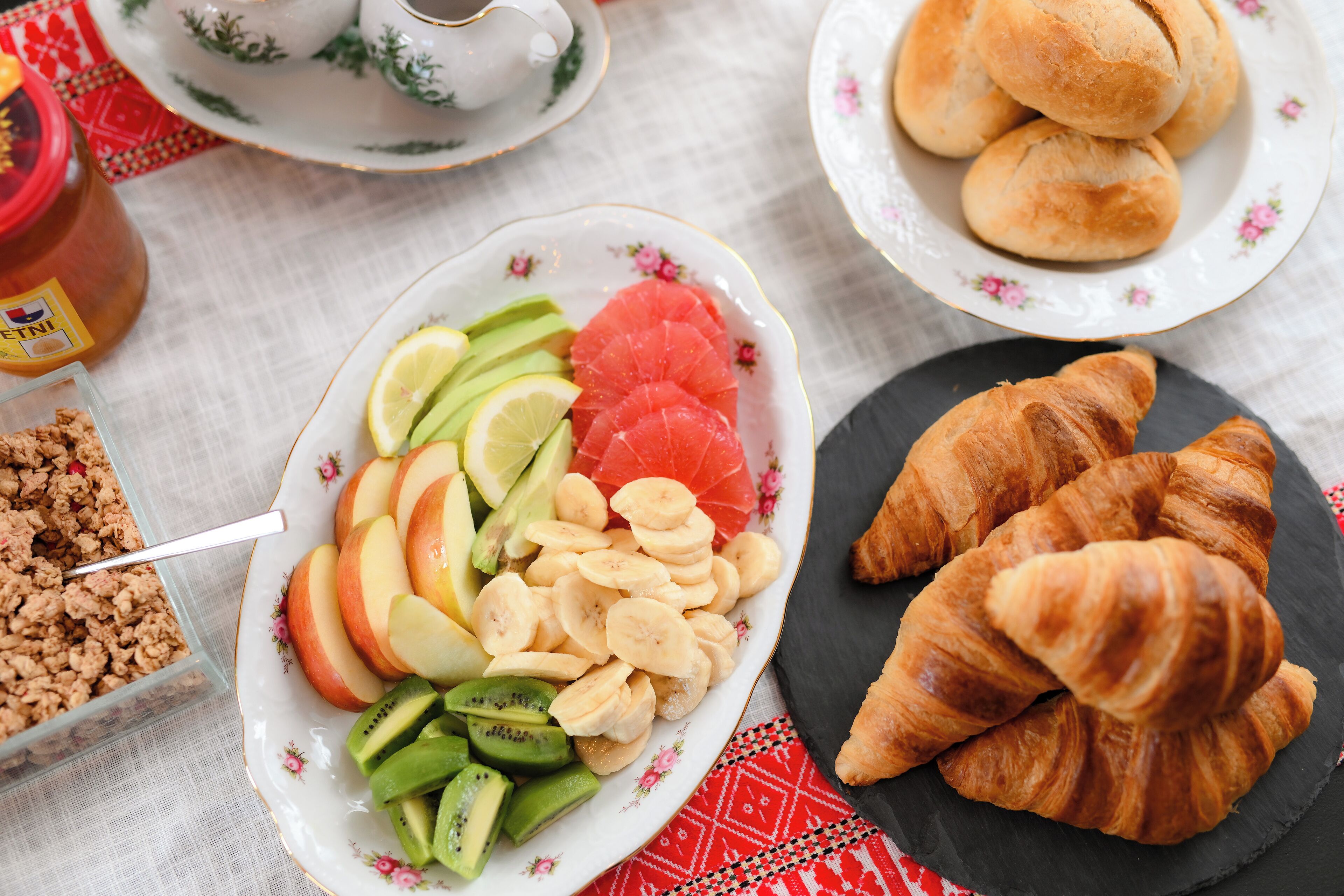 Una escena de desayuno con un plato de frutas y nueces en rodajas, un plato de croissants y un frasco de mermeladas.