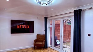 Une pièce dotée d’un grand téléviseur à écran plat affichant le logo « NETFLIX », d’un fauteuil en cuir marron, d’un plancher en bois et d’une porte-fenêtre coulissante donnant sur un espace extérieur.