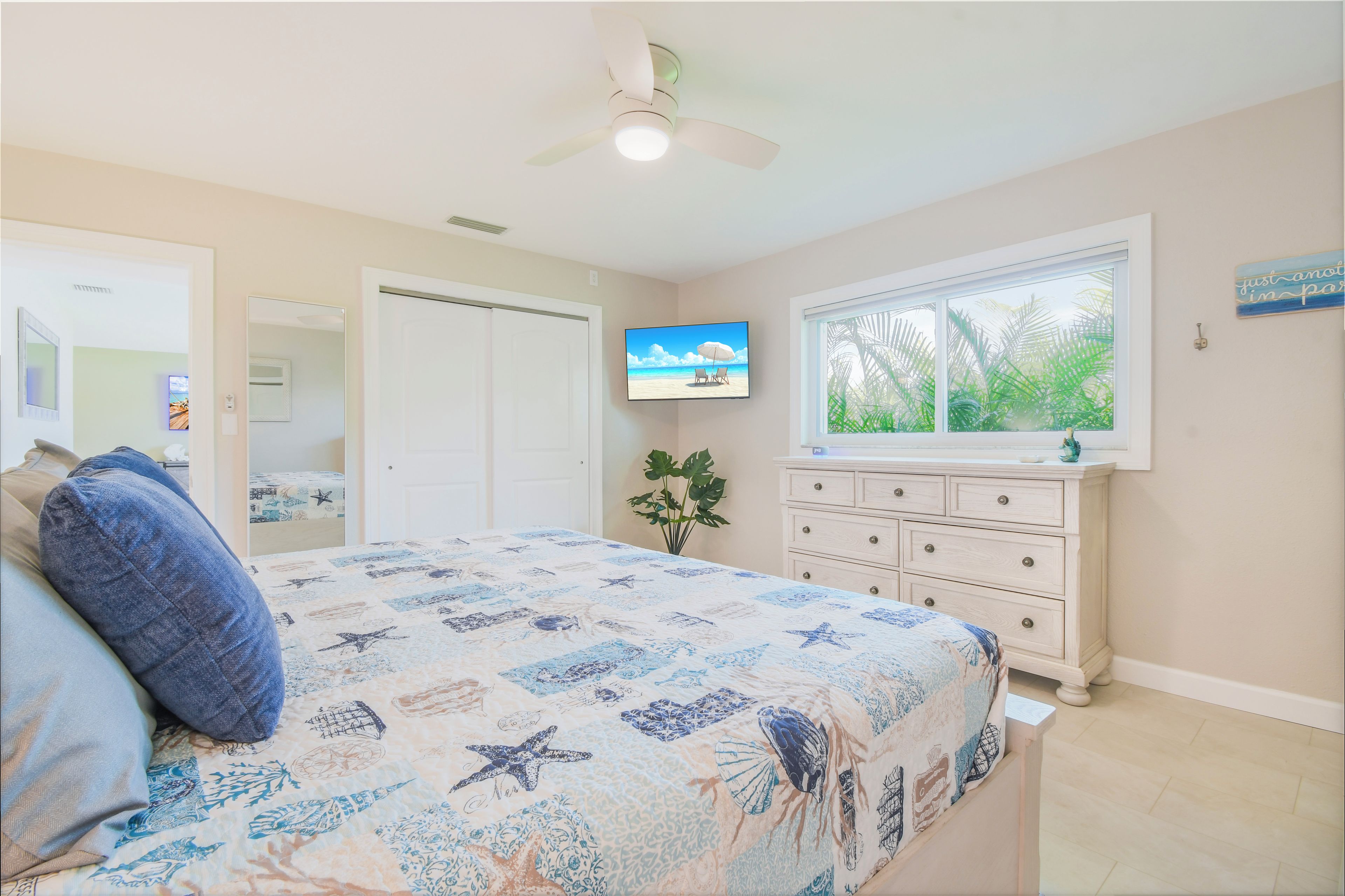 A bedroom with a bed, a dresser, a television, and a window with a view of palm trees.