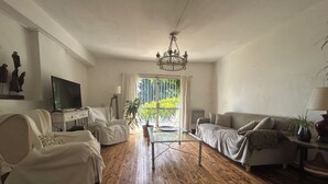 A living room with a wooden floor, white sofas, a glass coffee table, a TV, and a chandelier.