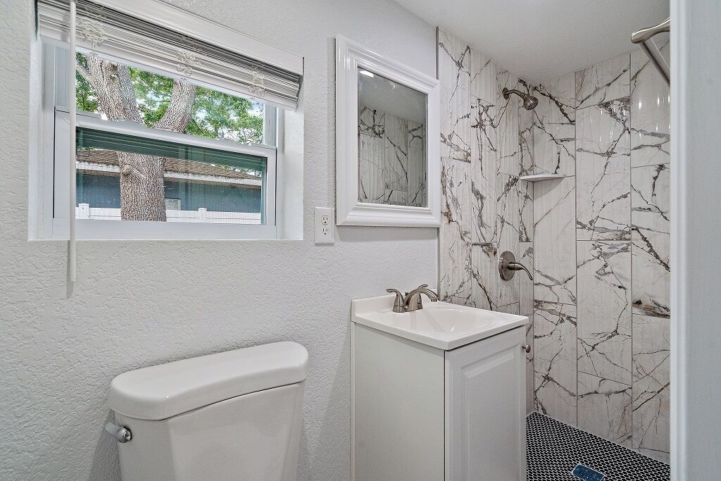 Un baño con inodoro, lavamanos y ducha. Hay una ventana sobre el inodoro y un espejo sobre el lavamanos.