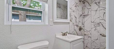 Un baño con inodoro, lavamanos y ducha. Hay una ventana sobre el inodoro y un espejo sobre el lavamanos.