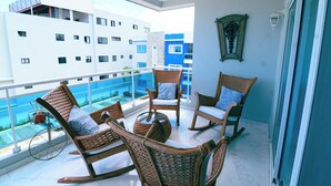 A balcony with wicker furniture, a view of a pool, and a building with blue and white facades. - Bellamoza, 3 bedrooms apartment. Peaceful, Airy and Beautiful views (Puerto Plata)