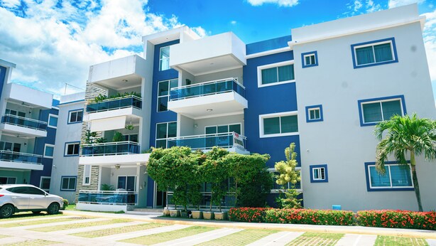 Un edificio moderno de varios pisos con balcones, un coche estacionado adelante y un cielo azul despejado.