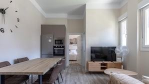 A modern kitchen with a refrigerator, oven, and microwave. There is a dining area with a wooden table and chairs, and a living area with a TV and fan. - Hospital Cruces Metro Bec (Barakaldo)
