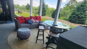 A covered outdoor seating area with a sofa, ottoman, and table, a barbecue grill, and a view of the surrounding greenery.
