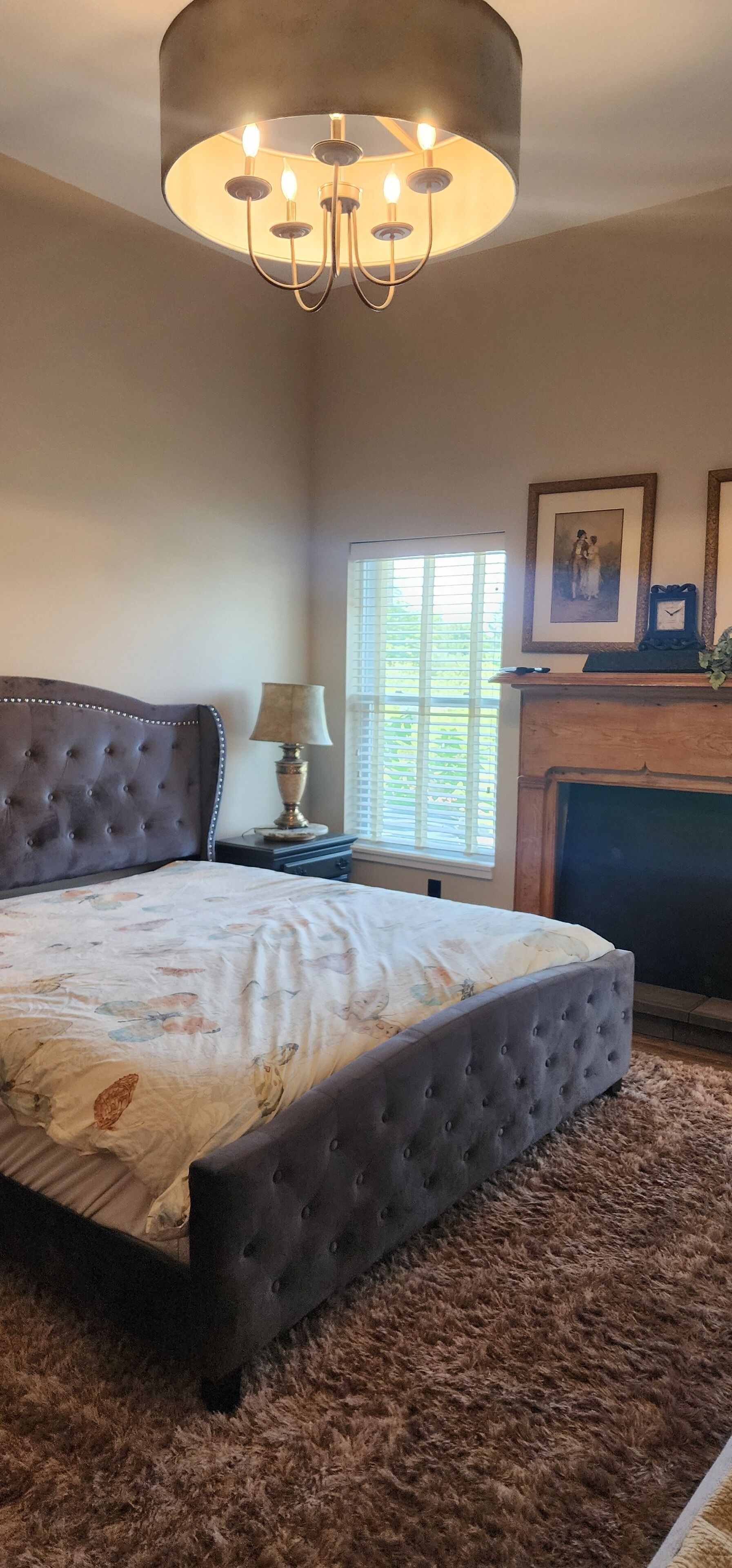 A bedroom with a tufted headboard, a large bed with a floral bedspread, a round table with a lamp, a fireplace, and a framed picture above it.