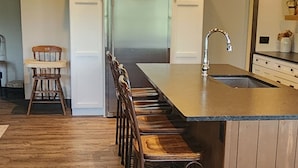 A kitchen with a wooden floor, a long island with stools, pendant lights, and a refrigerator.