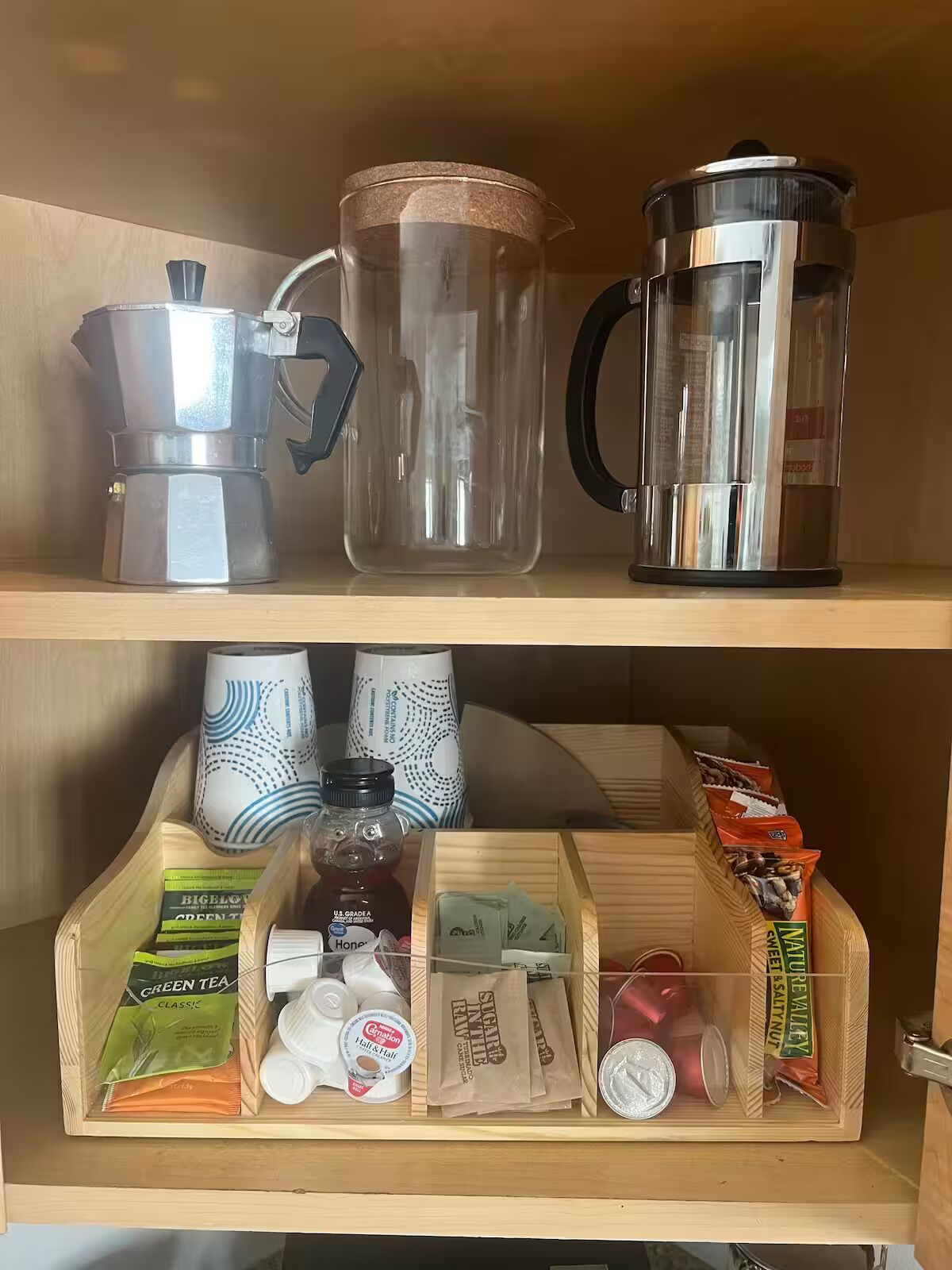 Una repisa de cocina con una cafetera, un jarro de vidrio y una olla de metal. Abajo, hay bolsitas de té, un tarro de miel y una caja de bocadillos.