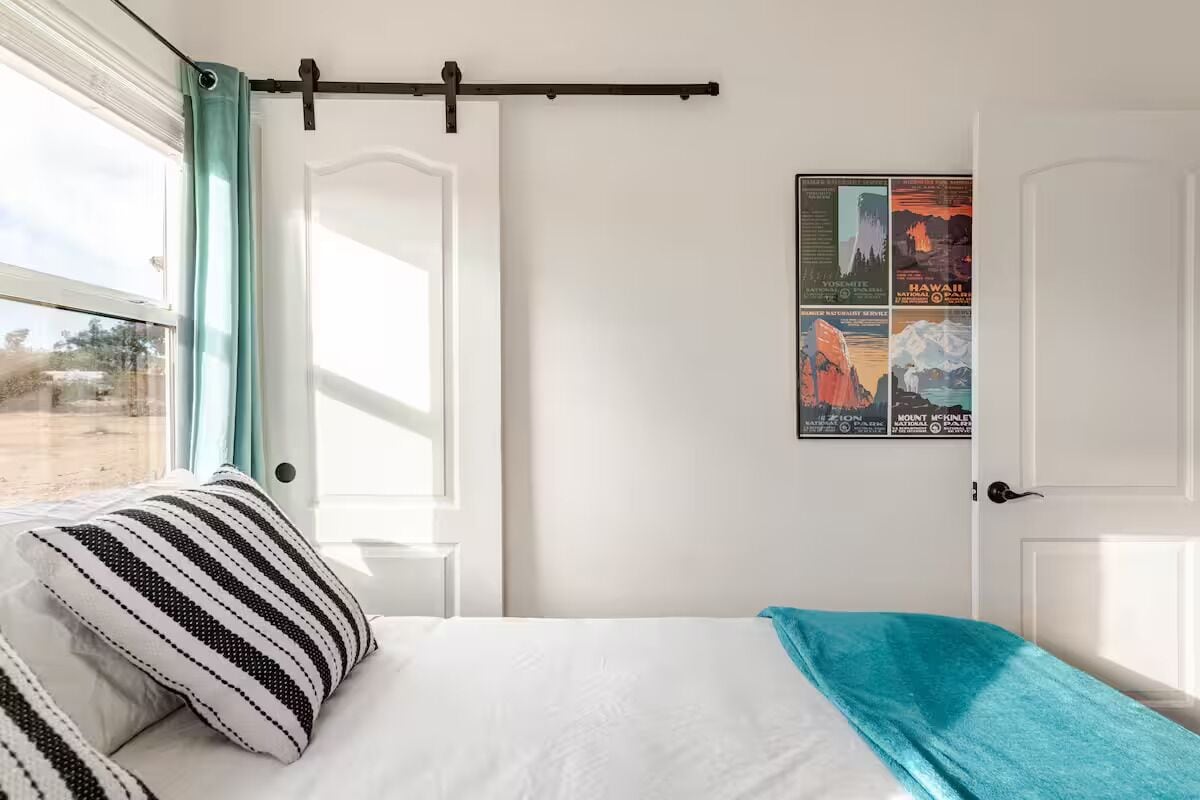 A hotel room with a bed, a window with curtains, a wall-mounted rack, and a poster with travel destinations.