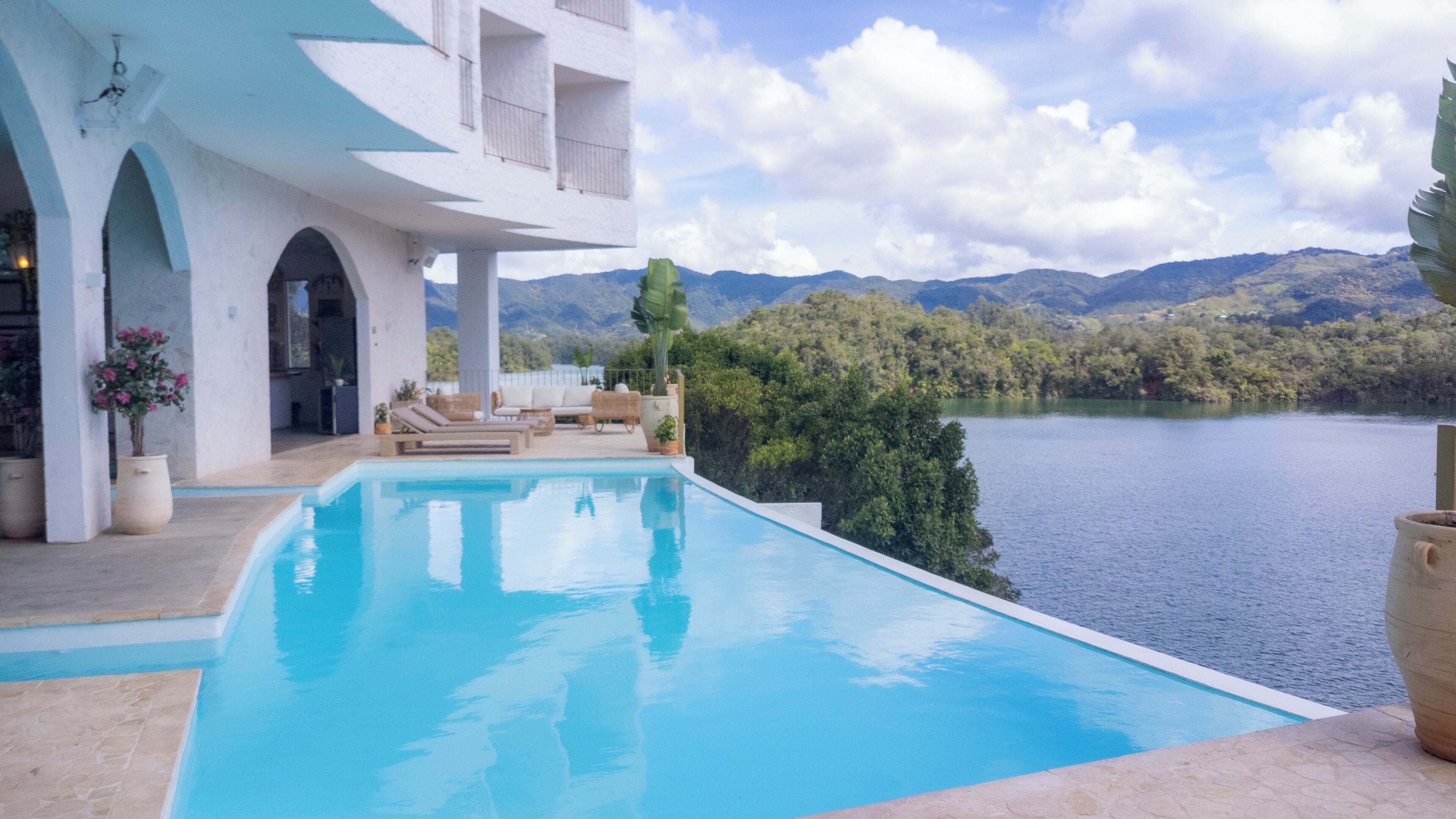 A pool with a view of mountains and a lake, a white building with balconies, and outdoor furniture.