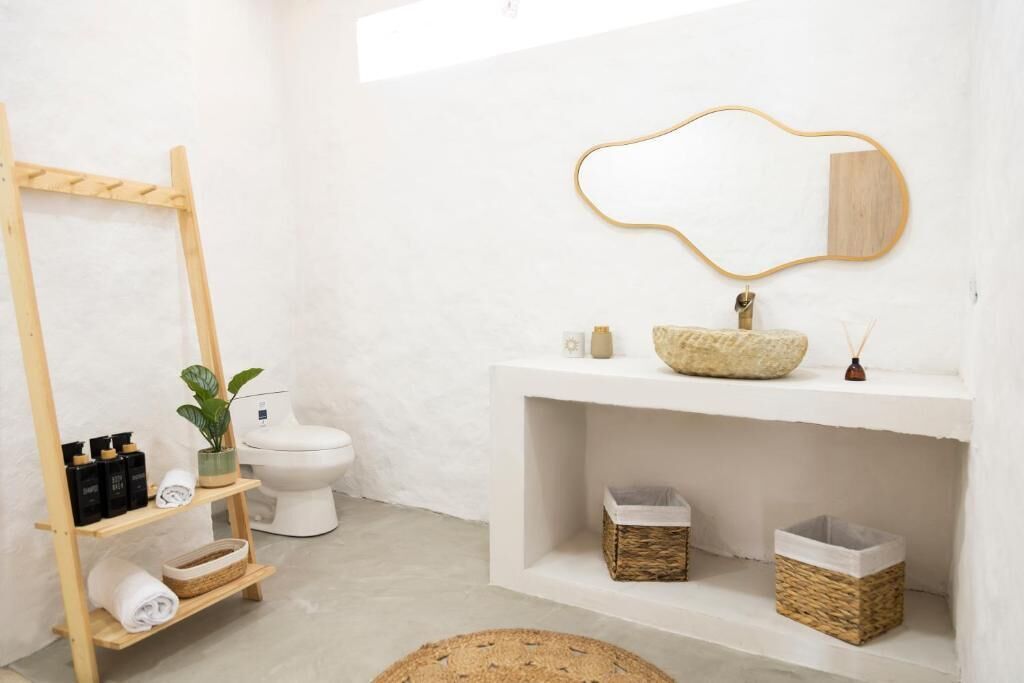 A minimalist bathroom with a wooden shelf holding toiletries, a white toilet, a large mirror, and a woven basket.