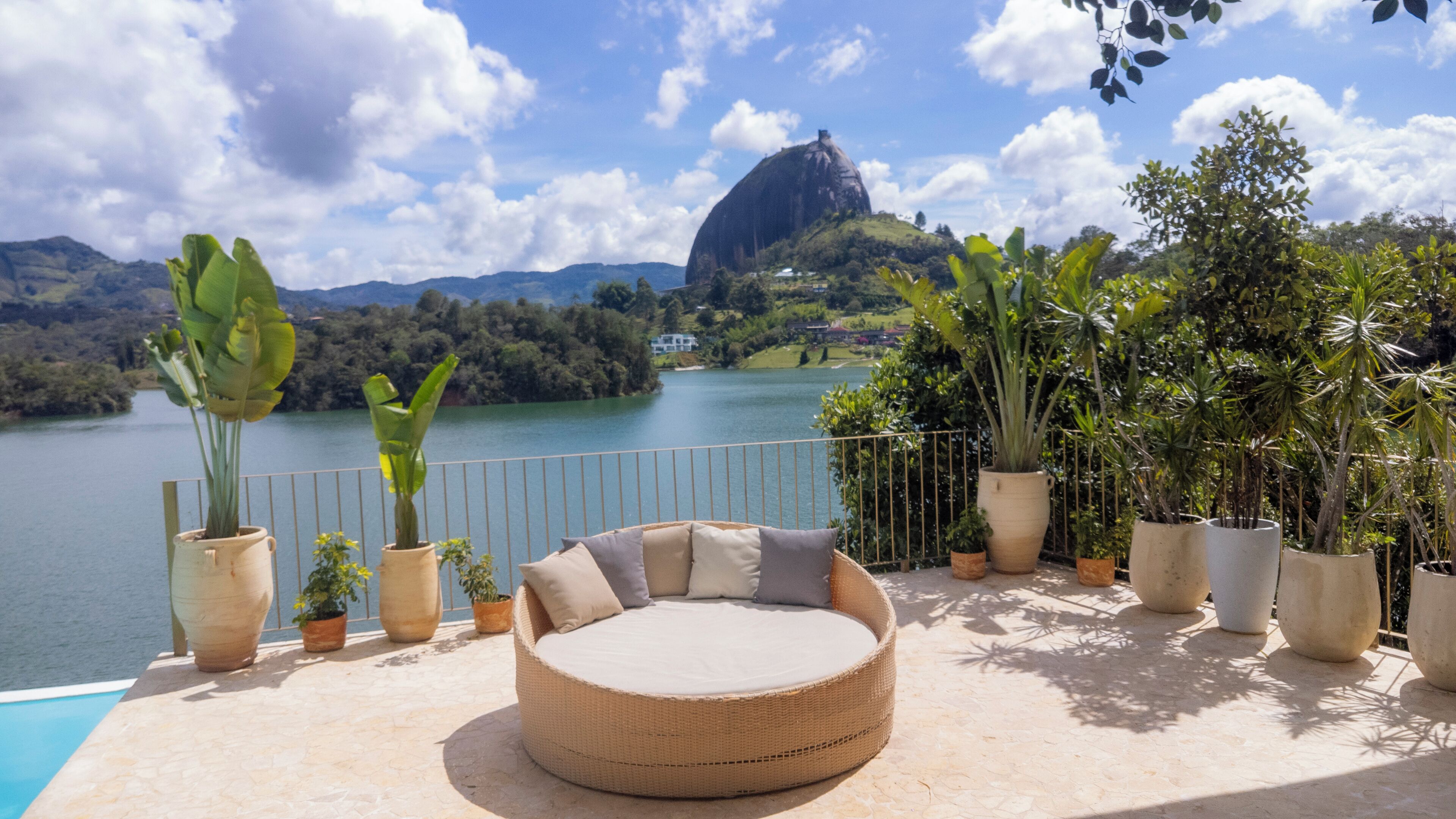 A round wicker sofa with cushions, potted plants, and a view of a lake with mountains in the background.
