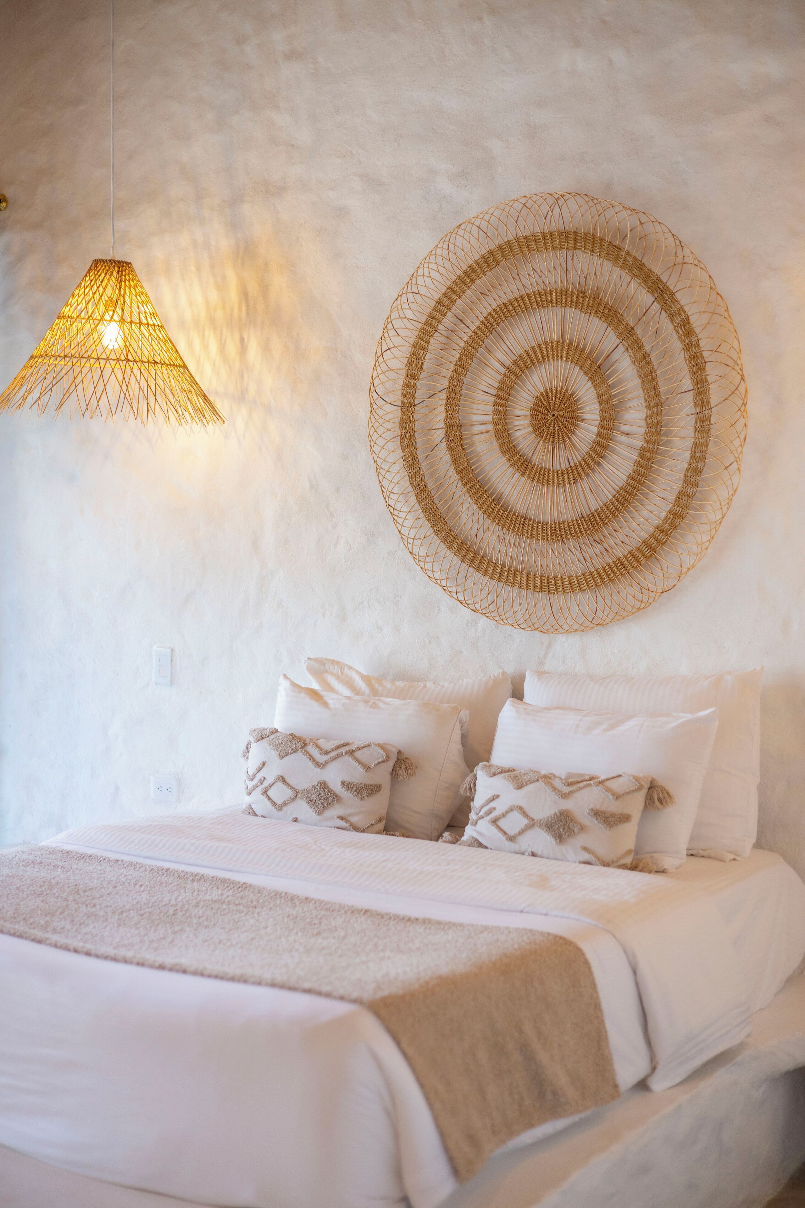A bed with white bedding, a woven wall hanging, and a hanging light fixture.