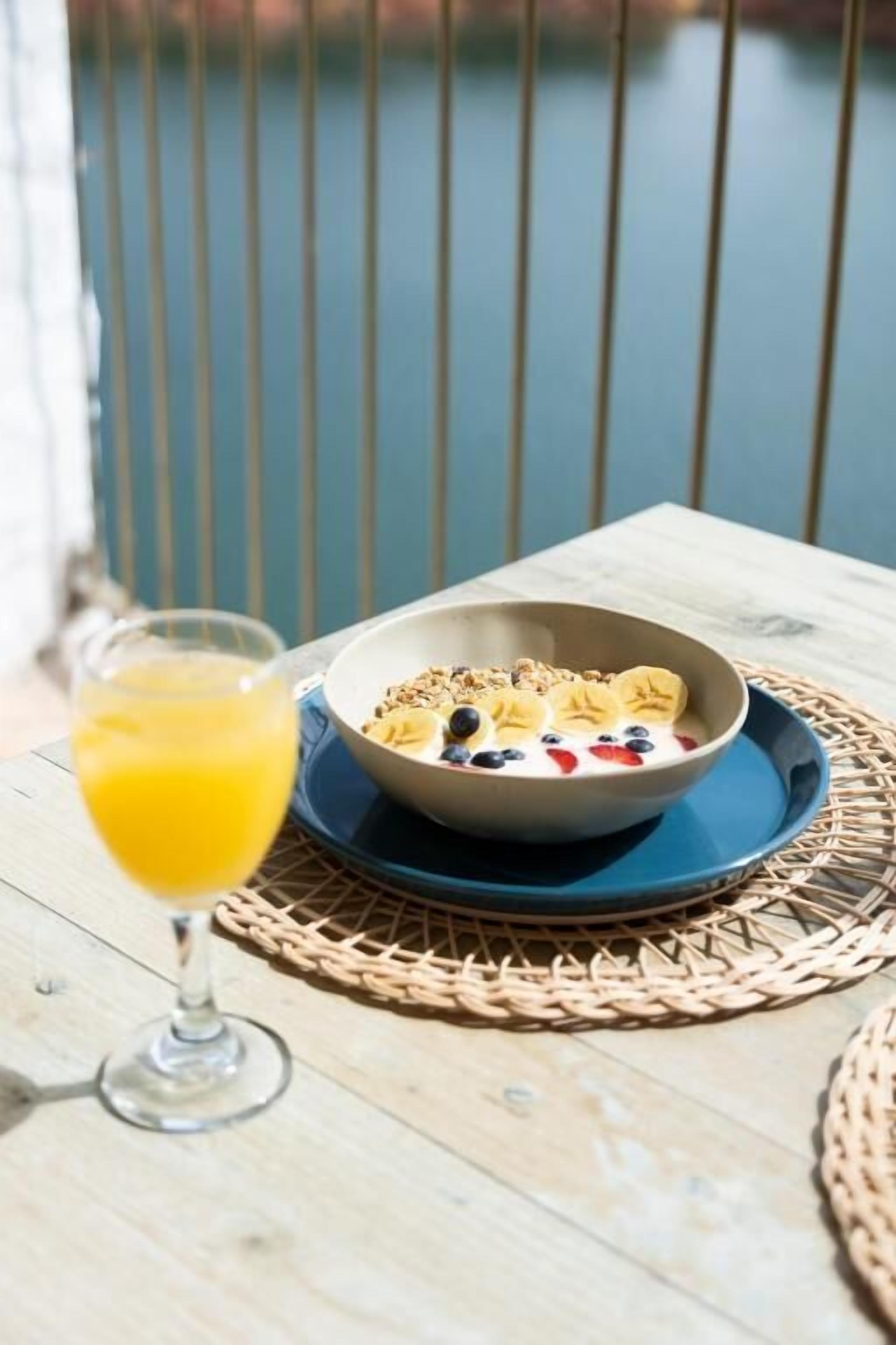 A breakfast setting with a bowl of cereal, fruits, and a glass of orange juice on a wooden table by a railing.