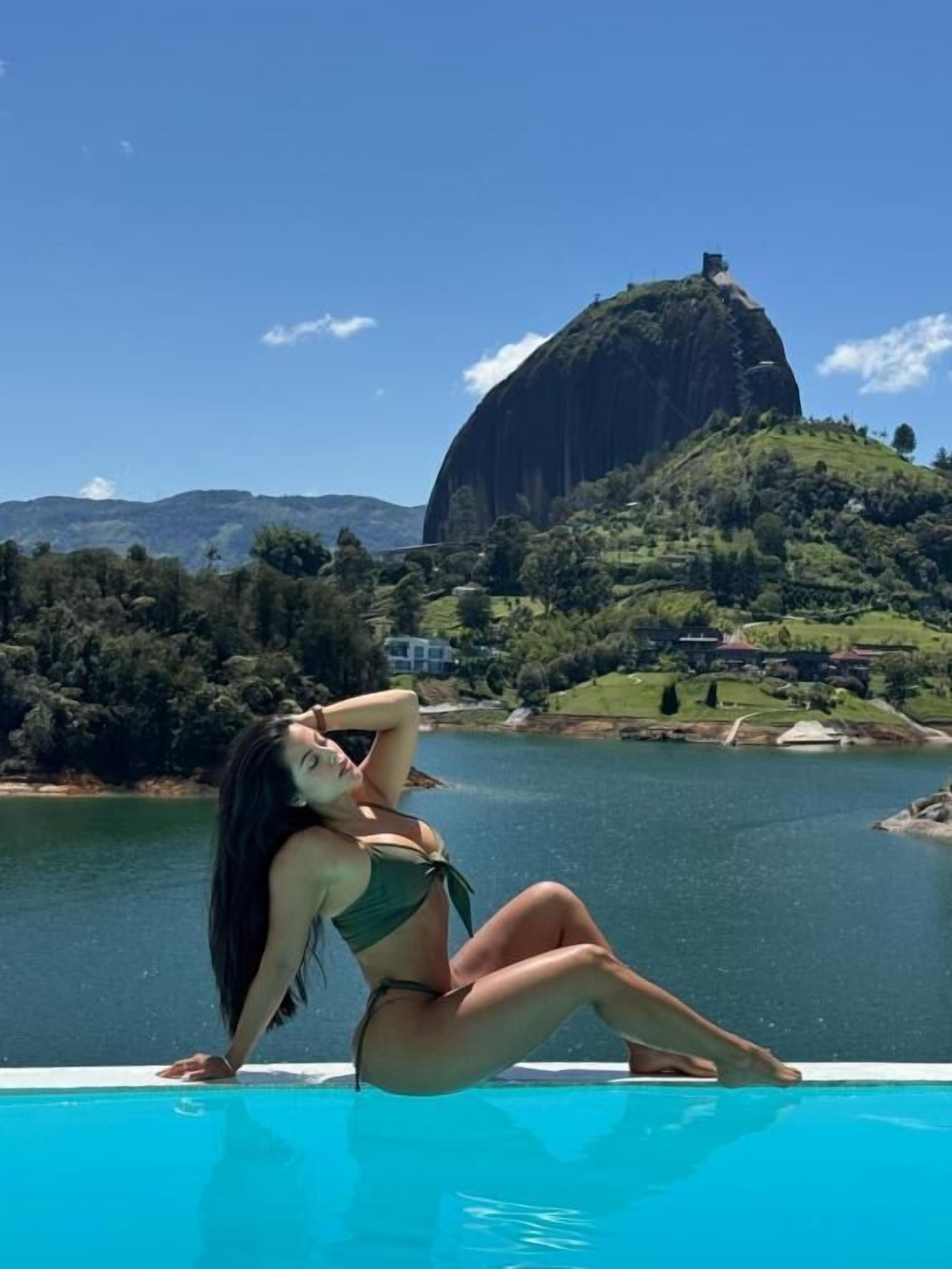A woman sitting by a pool with a large rock formation and hills in the background.