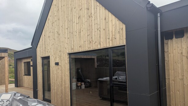 Ein modernes Gebäude mit Holzfassade und großen Glasüren, die zu einem Außenbereich mit Whirlpool und Grill führen.