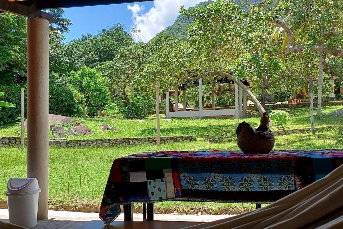 Un hamac sous une pergola, avec une table recouverte d’une nappe à motifs, un pot décoratif et une poubelle.