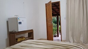 A room with a bed, a wooden nightstand, a white refrigerator, and an open door leading to a covered outdoor area. - Sitio Pedagógico Vale da Serra (Maranguape)