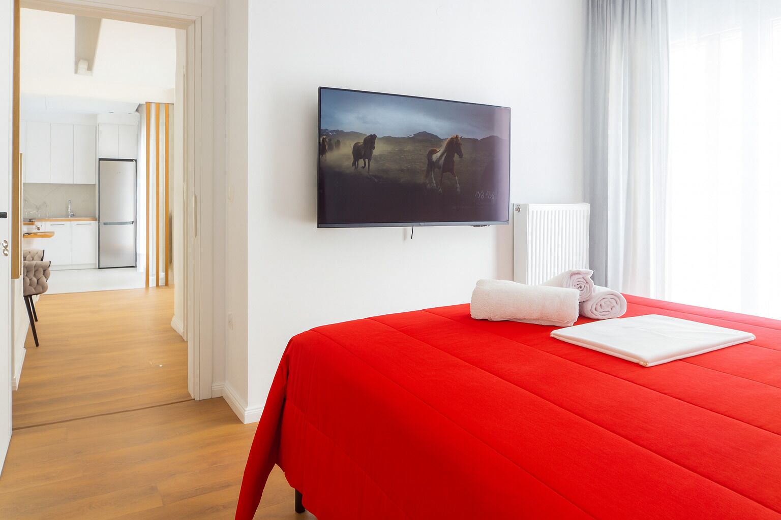 A hotel room with a red bedspread, a flat-screen TV mounted on the wall, and a view of a kitchen and dining area.
