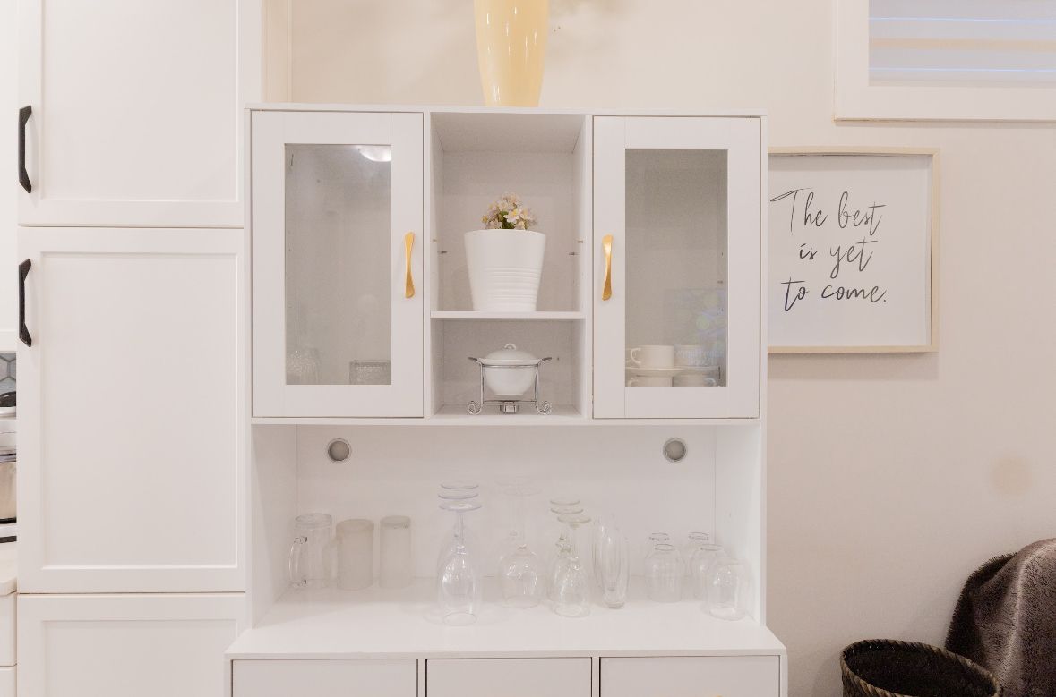 A white cabinet with glass doors, a white vase with flowers, and a framed quote on the wall.