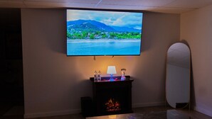 A room with a flat-screen TV mounted on the wall displaying a scenic landscape, a small table with a lamp and decorative items, a fireplace, and a mirror.