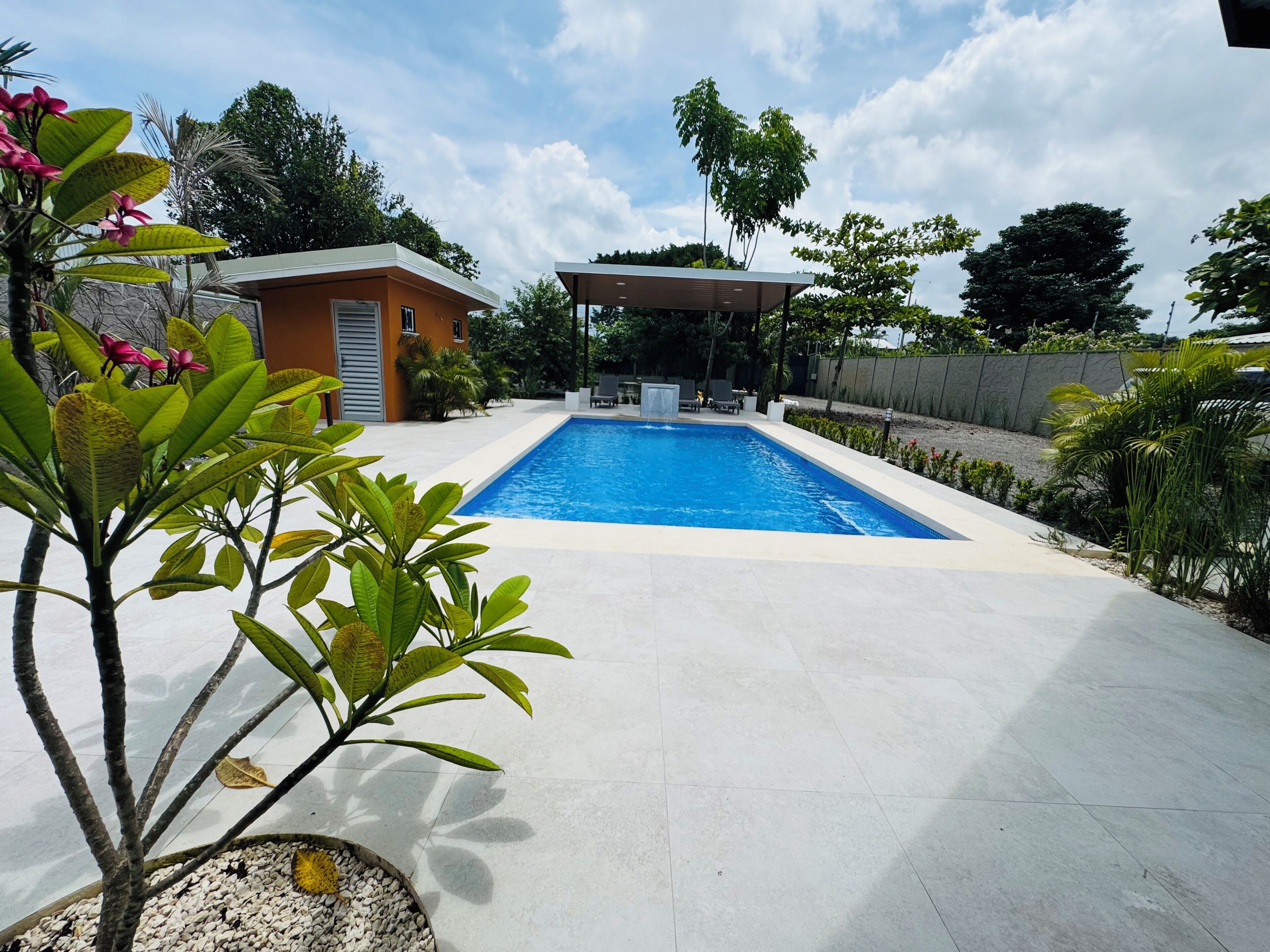 Une piscine rectangulaire entourée d’une terrasse carrelée, un espace de détente couvert et un petit bâtiment doté d’une porte et de fenêtres.
