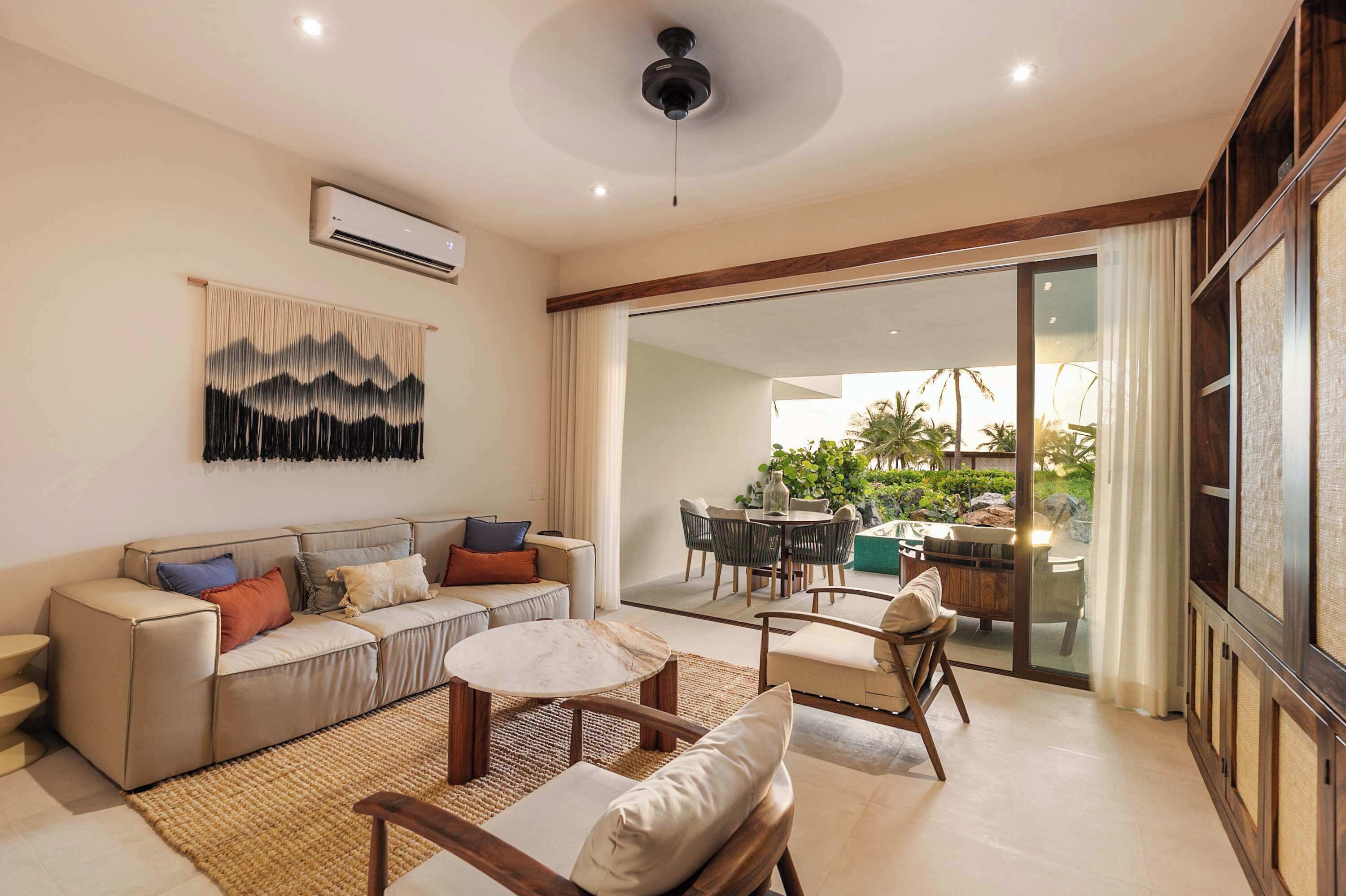 Una sala de estar moderna con sofá, mesa de centro y sillas, un ventanal con vista a un jardín, y un equipo de aire acondicionado en la pared.
