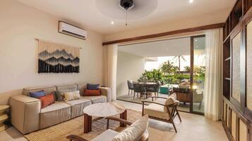 Una sala de estar moderna con sofá, mesa de centro y sillas, un ventanal con vista a un jardín, y un equipo de aire acondicionado en la pared.