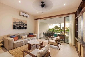 Un salon moderne avec un canapé, une table basse et des fauteuils, une grande fenêtre donnant sur un jardin et un appareil de climatisation fixé au mur.