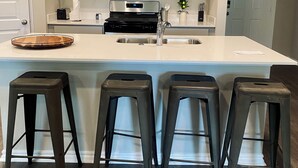 A modern kitchen with a central island, four stools, stainless steel appliances, and open shelving. - Brand New home, 4 beds Bison Ridge Retreat Ft Sill (Lawton)
