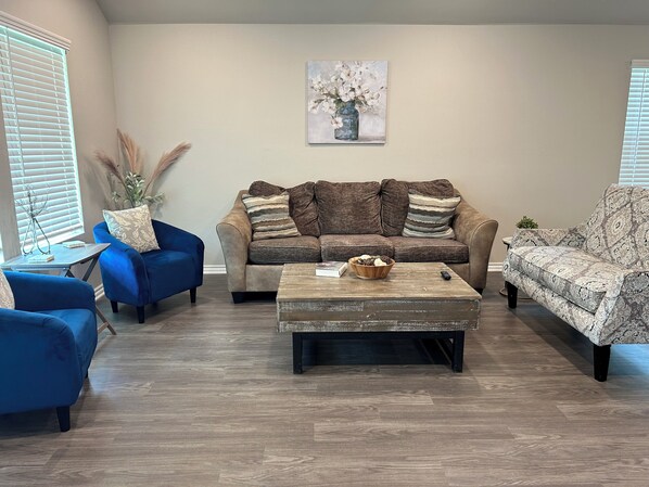 A living room with a brown sofa, blue armchairs, and a patterned armchair. A wooden coffee table with a bowl and remote control is in the center. - Brand New home, 4 beds Bison Ridge Retreat Ft Sill (Lawton)