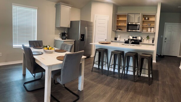 A modern kitchen with a dining area, featuring a white table, grey chairs, and a central island with bar stools. - Brand New home, 4 beds Bison Ridge Retreat Ft Sill (Lawton)
