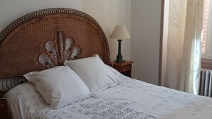 Een bed met een rieten hoofdbord, twee bedlampen en een raam met zonwering.