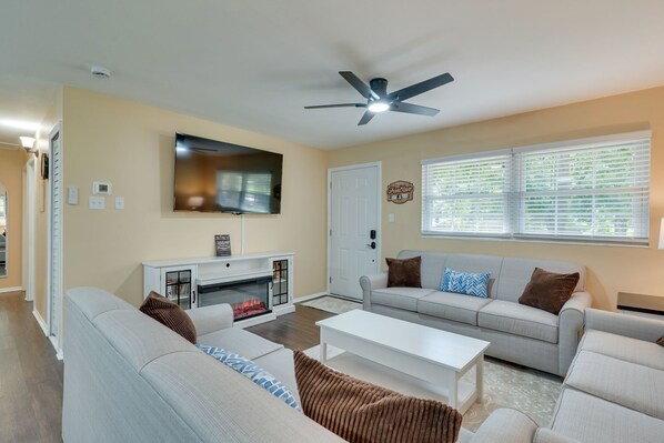 A living room with a fireplace, a ceiling fan, and a large window with blinds.