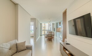 A modern living room with a sofa, a flat-screen TV mounted on the wall, and a dining area in the background. - Apartments in the Turquesa Building - Balneario Picarras - RTS (Picarras)
