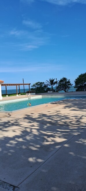 A clear sky, a swimming pool, and a beach area with trees and a building in the background. - Charming 1 Bedroom Condo in peaceful, secure, gated community with pool (Portmore)