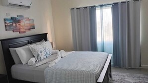 Una cama bien hecha con cabecera, aire acondicionado de pared y una ventana con cortinas.