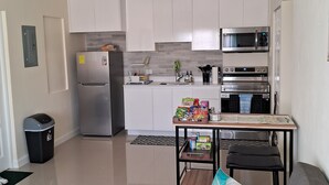 Una cocina moderna con armarios blancos, un refrigerador de acero inoxidable, un microondas y una estufa. Hay una pequeña mesa de comedor con sillas y un cubo de basura cerca de la cocina.