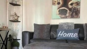 Un salon avec un canapé gris, un coussin orné du mot « Home » et un tableau sur le thème des vaches accroché au mur.