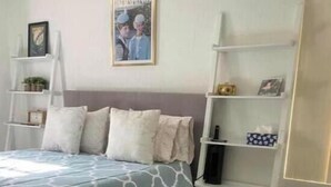Une chambre à coucher avec un lit, une couvre-lit grise à motifs, un tapis gris et blanc, une étagère blanche contenant divers objets et un tableau encadré accroché au mur.