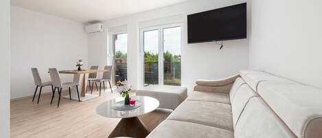 Uma sala de estar moderna com sofá, mesa de jantar com cadeiras, TV fixada na parede e varanda com porta de vidro.