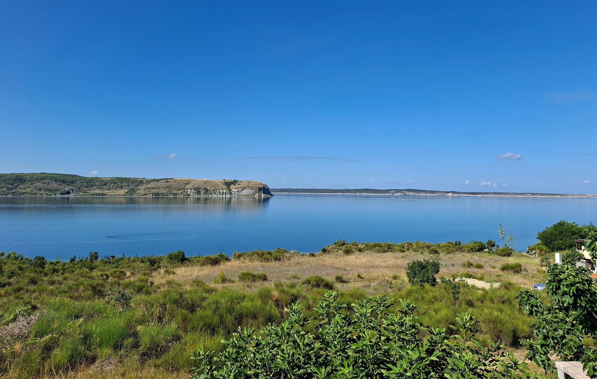 A coastal landscape with a calm body of water, a distant hill, and a clear sky.