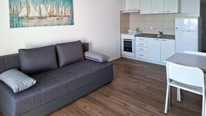 A modern living room with a grey sofa, a white dining table, and a kitchen area with white cabinets and a built-in oven. - Lovely apartment in Rtina with WiFi (Rtina)