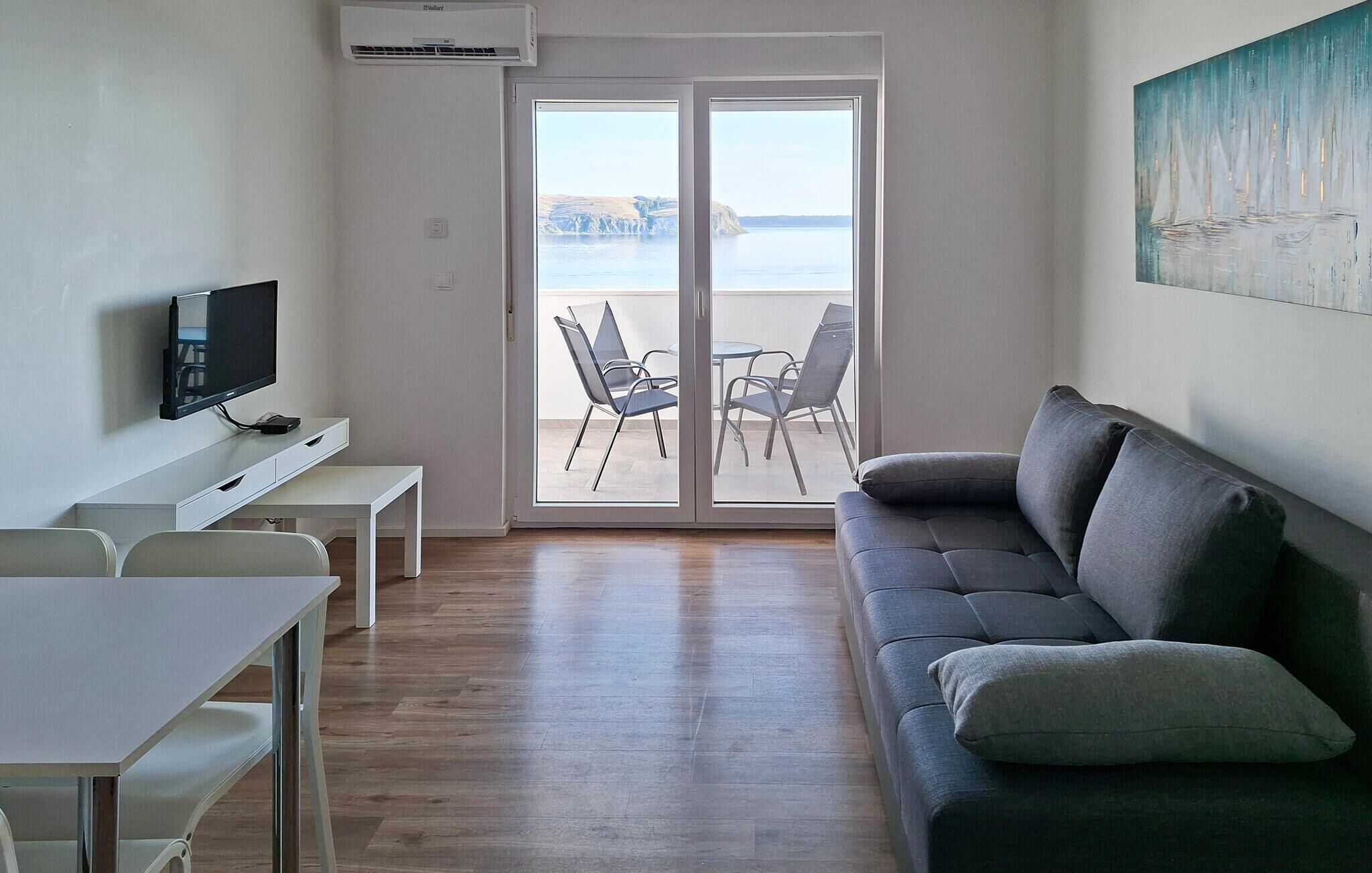 Sala de estar moderna com televisão de tela plana, sofá, mesa de jantar e cadeiras. Há uma porta de vidro deslizante que dá para uma área externa com vista para o mar e falésias.